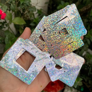 Puede incluir: Un conjunto de tarjetas de pendientes cuadradas e iridiscentes con un recorte cuadrado central y pequeños agujeros para los pendientes. Las tarjetas tienen un efecto brillante y con purpurina, que refleja varios colores. Las tarjetas se sostienen en una mano sobre un fondo verde borroso.