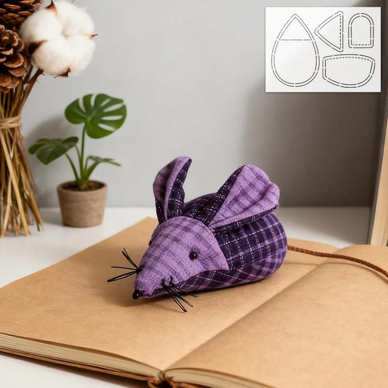 May include: A handcrafted fabric mouse in purple and black plaid, with black whiskers and a long tail. The mouse is positioned on an open book. A pattern template is visible in the background.