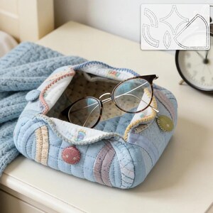 May include: A handmade, quilted fabric box in shades of blue, with a patchwork design and decorative buttons. Tortoiseshell eyeglasses rest inside the open box. A blue blanket is visible in the background. A pattern template is in the background.