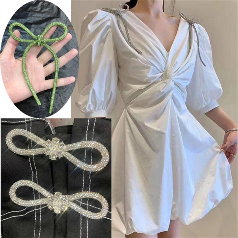 May include: The image showcases a white dress with a V-neck and puff sleeves, adorned with sparkling embellishments. A green, rhinestone-covered bow and two silver, rhinestone-encrusted closures are also featured, offering a touch of glamour.