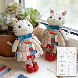Può includere: Due bambole gatto in tessuto fatte a mano con tratti del viso ricamati e occhi blu. Ogni gatto indossa un vestito patchwork, un fiocco blu al collo e stivali rossi. Una sagoma in plastica bianca con forme per il taglio è in primo piano.