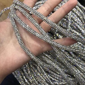 May include: Close-up of several strands of sparkling silver rhinestone trim. The trim is densely covered in small, reflective rhinestones, creating a shimmering effect. The strands are held in a hand, with more trim visible in the background.
