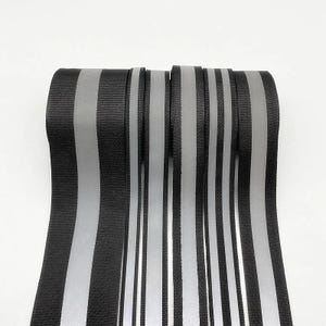 Puede incluir: Primer plano de cinta reflectante negra y plateada. La cinta tiene rayas verticales negras y plateadas, creando un patrón de alto contraste. El material parece estar tejido y la cinta está enrollada, mostrando los bordes.