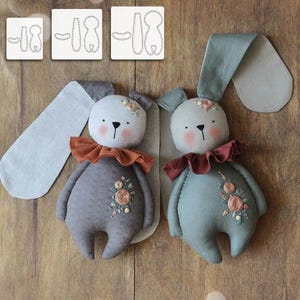 May include: Two handmade bunny dolls in gray and blue-green with floral embroidery and ruffled collars. The bunnies have long, floppy ears and are next to three white templates with bunny outlines.