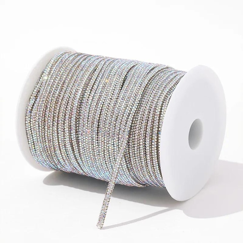 May include: A spool of shimmering rhinestone chain. The chain is tightly wound around a white plastic spool. The rhinestones are clear and reflect light, creating a sparkling effect. The chain is ideal for jewelry making and craft projects.