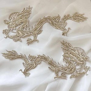 Puede incluir: Dos apliques bordados dorados con forma de dragón. Los dragones están detallados con costuras intrincadas, escamas, garras y cuerpos fluidos. Los apliques se colocan sobre un fondo blanco ligeramente texturizado.