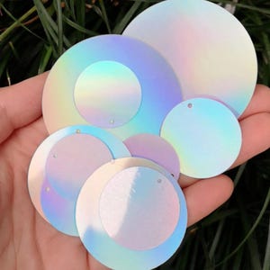 Puede incluir: Una colección de lentejuelas circulares iridiscentes de varios tamaños. Las lentejuelas tienen un agujero para ensartar y muestran un efecto de cambio de color con tonos de azul, morado y amarillo. Las lentejuelas se sostienen en una mano.