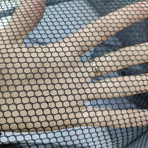 Puede incluir: Una tela de malla negra con un patrón hexagonal consistente. El material es semitransparente, lo que permite una visión clara de la superficie debajo de la estructura de rejilla. El tejido es uniforme, creando un diseño geométrico de panal.