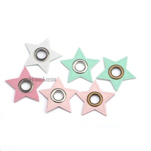 Op de afbeelding: Zes stervormige stoffen patches in pastelkleuren, waaronder lichtroze, mintgroen en zilver. Elke ster is voorzien van een centrale metalen ring, met een zilverkleurige of bronskleurige afwerking. Decoratieve stiksels omlijnen de randen van de sterren.