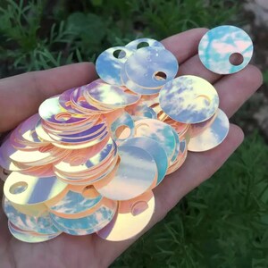 Peut inclure: Une collection de sequins circulaires iridescents présentant un motif de ciel imprimé avec des nuages blancs sur fond bleu clair. Chaque disque plat et rond possède un petit trou percé près du bord, créant une finition brillante et réfléchissante.