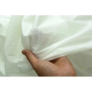 Op de afbeelding: Close-up van een hand die een stuk witte stof vasthoudt. De stof lijkt licht en enigszins doorschijnend te zijn, met een subtiele glans. Het materiaal is geplooid en gedrapeerd, wat de textuur en de plooien van de stof laat zien.