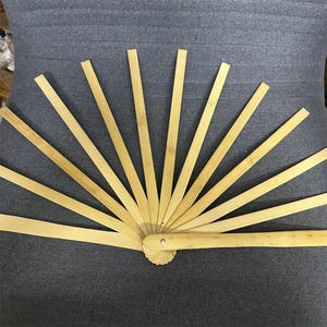 Può includere: Un ventaglio a mano in bambù marrone chiaro. Il ventaglio è parzialmente aperto, mostrando sottili stecche rettangolari in bambù che si irradiano da un punto centrale. La base del ventaglio presenta un design decorativo intagliato. Il ventaglio è appoggiato su una superficie grigia.