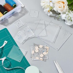 Puede incluir: Un conjunto de plantillas de acolchado de acrílico transparente en varios tamaños rectangulares y cuadrados, con orificios preperforados. El set incluye una pieza de tela gris con un patrón floral blanco, junto a carretes de hilo y tijeras.