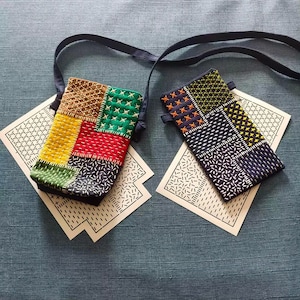 May include: Two rectangular crossbody pouches featuring intricate sashiko-style embroidery. One pouch displays a vibrant patchwork of yellow, red, green, and brown fabric squares. The second pouch features a dark blue base with yellow and orange stitched patterns.
