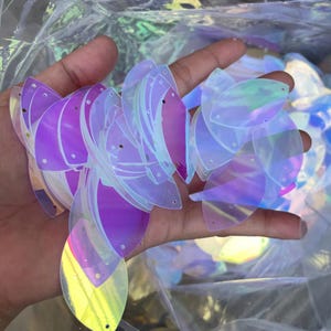 Peut inclure: Une collection de sequins iridescents en forme de feuille, dotés d'une surface lisse et réfléchissante avec une finition changeante. Chaque sequin possède deux petits trous pré-percés en haut et en bas pour la fixation, affichant des teintes violettes, bleues et jaunes.
