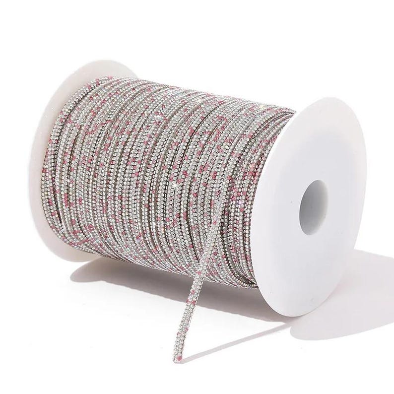 May include: A spool of silver rhinestone chain with small pink accents. The chain is tightly wound on a white plastic spool. The rhinestones are small and closely set, creating a sparkling effect. Ideal for jewelry making and crafts.