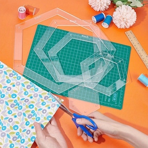 May include: Clear acrylic quilting templates, sized from 5 cm to 15 cm, are arranged on a green cutting mat. A pair of blue-handled scissors cuts floral patterned fabric. Spools of thread and a wooden ruler are also present.