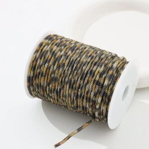 May include: A spool of decorative cord with a camouflage pattern in shades of gray, brown, and gold. The cord is tightly wound on a white plastic spool. The end of the cord is visible, showing the multi-colored strands.