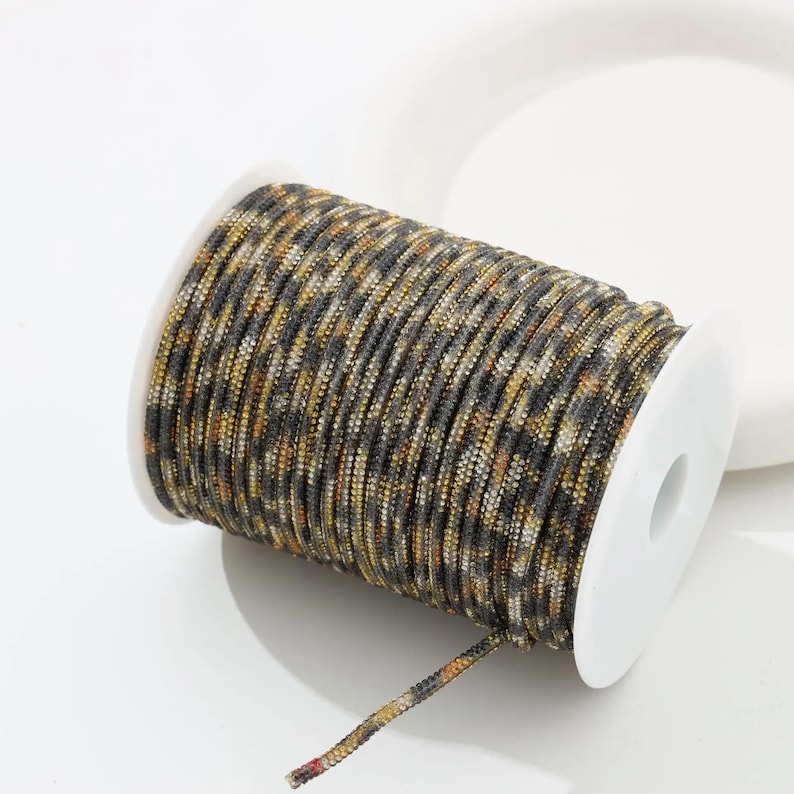 May include: A spool of decorative cord with a camouflage pattern in shades of gray, brown, and gold. The cord is tightly wound on a white plastic spool. The end of the cord is visible, showing the multi-colored strands.