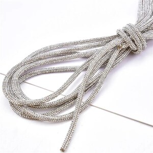 May include: Silver rhinestone trim, knotted at the top, lies on a white surface. The trim is made of multiple strands of small, sparkling rhinestones, creating a shimmering effect. The strands are looped and arranged in a decorative pattern.