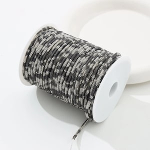 May include: A spool of camouflage patterned cord in shades of gray and black. The cord is tightly wound around a white plastic spool. The cord has a textured appearance and is suitable for various crafting projects.