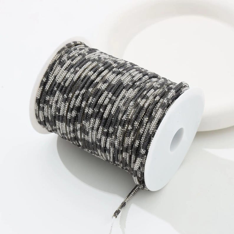 May include: A spool of camouflage patterned cord in shades of gray and black. The cord is tightly wound around a white plastic spool. The cord has a textured appearance and is suitable for various crafting projects.