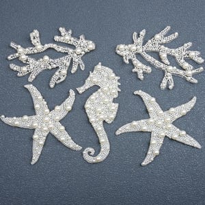 May include: A collection of five decorative appliqués featuring two coral branches, two starfish, and one seahorse. Each piece is intricately covered in small, white, round beads and pearls, creating a textured, uniform surface across all shapes.