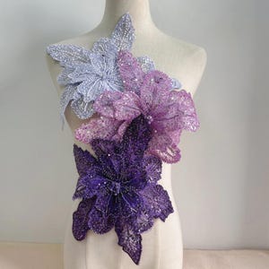 May include: A layered floral applique featuring three distinct flower shapes in shades of light lavender, medium purple, and deep violet. Each petal is densely covered in shimmering sequins and intricate beadwork, creating a textured, dimensional surface.