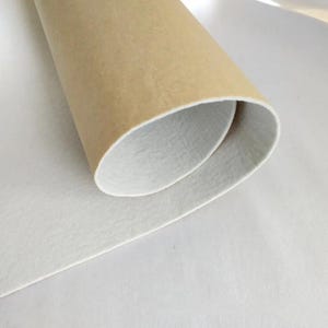 Puede incluir: Una hoja enrollada de material de color beige con un interior blanco, sobre una superficie blanca. El material beige tiene una textura ligeramente rugosa, mientras que el interior y la superficie son lisos. La imagen es un primer plano del material enrollado.
