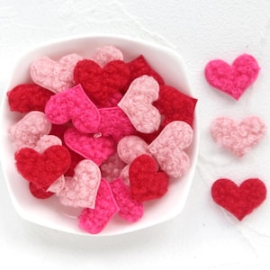 Puede incluir: Un cuenco blanco lleno de corazones de fieltro surtidos en tonos de rojo, rosa y rosa claro. Tres corazones adicionales están dispuestos a la derecha. Los corazones tienen un aspecto texturizado y difuso.