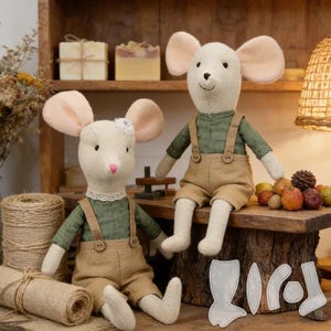 May include: Two stuffed mouse dolls made of cream-coloured fabric with large pink-lined ears. Each mouse wears a green long-sleeved shirt and tan overalls with buttons. One mouse has a white lace collar and a small flower detail on its head.