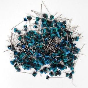 May include: A pile of small, decorative feathers with teal, blue, and black coloring. Each feather is attached to a thin, silver-colored wire. The feathers are scattered on a white background.