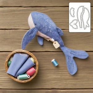 May include: A handmade blue whale plush toy with a cream-colored belly and fins. The whale has embroidered details and a small tag attached. A basket of fabric scraps and spools of thread are nearby, along with a pattern.