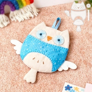 Peut inclure: Une pochette en feutre en forme de hibou faite à la main, de couleur crème et bleue. Le hibou a de grands yeux bleus, un bec orange et des ailes blanches. La section bleue présente un motif de gouttes de pluie. Une boucle bleue est attachée pour suspendre.