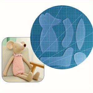 May include: A plush mouse doll with a pink dress and a ruffled collar sits next to a set of clear plastic sewing templates. The templates are in the shape of the mouse doll and its parts, such as the ears and feet.