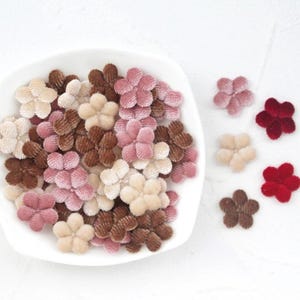 Può includere: Una collezione di piccoli abbellimenti floreali in velluto in varie tonalità di rosa, marrone e crema. Alcuni fiori sono disposti in una ciotola bianca, altri sparsi su una superficie bianca. Ideali per progetti di artigianato.