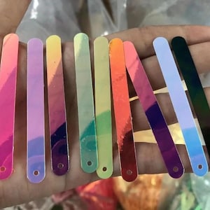 Puede incluir: Una colección de palitos artesanales rectangulares iridiscentes en varios colores, incluyendo rosa, morado, amarillo, verde, naranja y azul. Cada palito tiene una parte superior redondeada y un pequeño agujero en la parte inferior. Los palitos se sostienen en una mano.