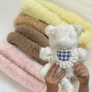 May include: A white plush teddy bear with a blue and white checkered bib and lace trim. The bear is held in front of a backdrop of fluffy, faux fur in shades of pink, brown, and yellow.