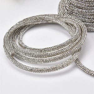 May include: Coiled silver rhinestone trim. The trim is made of small, sparkling rhinestones set closely together, creating a continuous, shimmering band. The trim is flexible and appears to be on a roll, suitable for various craft and design projects.