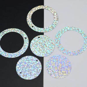 Puede incluir: Una colección de lentejuelas iridiscentes y brillantes de varias formas. La imagen presenta varias lentejuelas circulares y en forma de anillo, dispersas sobre un fondo blanco y negro. Las lentejuelas brillan con un arcoíris de colores.