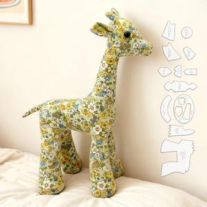 May include: A fabric giraffe toy with a floral pattern in yellow, blue, and white on a green background. The giraffe is standing on a white surface. Sewing pattern pieces are to the right.