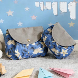 Puede incluir: Dos bolsas azules con estampado floral, solapas grises de imitación de ante y cierres de botón dorados. Las bolsas son de diferentes tamaños y están sobre una superficie gris. El estampado floral incluye flores blancas y crema.