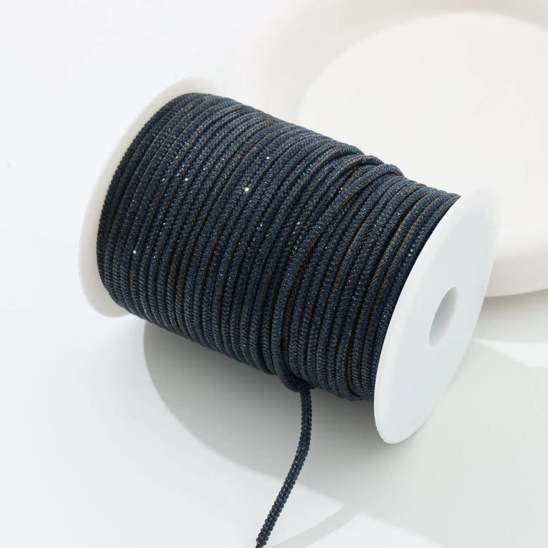 May include: A spool of dark blue, sparkly cord. The cord is tightly wound around a white plastic spool. The cord has a subtle sheen and is suitable for various crafting projects. The cord is a dark navy blue color.