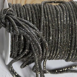 May include: A spool of dark gray, sparkly rhinestone trim. The trim is tightly wound on a white spool, with the sparkling rhinestones catching the light. This decorative trim is suitable for various craft projects.