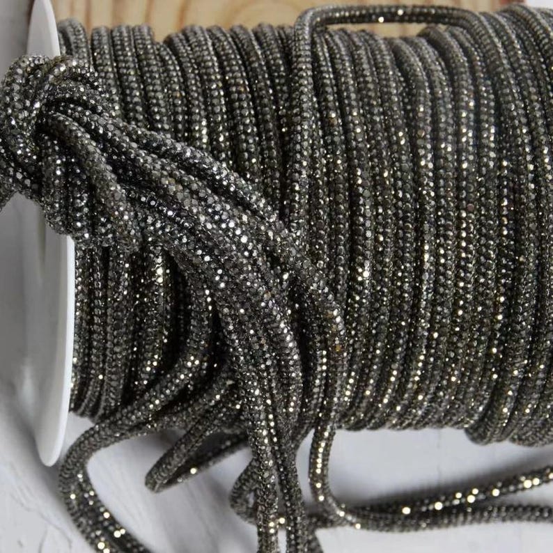 May include: A spool of dark gray, sparkly rhinestone trim. The trim is tightly wound on a white spool, with the sparkling rhinestones catching the light. This decorative trim is suitable for various craft projects.