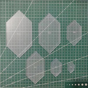 Puede incluir: Seis plantillas de acrílico transparente con forma hexagonal alargada, cada una con una etiqueta de tamaño impresa. Las plantillas están dispuestas en orden descendente de tamaño, de 15 cm a 2,5 cm, diseñadas para cortes y acolchados.