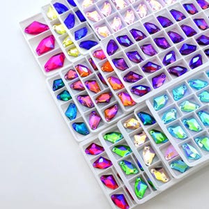 Può includere: Un assortimento di strass colorati e sfaccettati di varie forme e dimensioni, disposti in vassoi di plastica bianca. Gli strass mostrano una gamma di colori, tra cui rosa, viola, blu, verde e giallo, con una finitura iridescente. Ideale per l'artigianato e l'abbellimento.