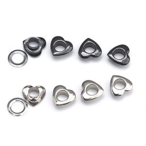 Könnte beinhalten: Eine Ansammlung von herzförmigen Metallösen und Unterlegscheiben, angeordnet in zwei Reihen. Die obere Reihe zeigt dunkelgraue, metallische Ösen, während die untere Reihe silberfarbene Ösen mit einer zentralen runden Öffnung für Stoffe präsentiert.