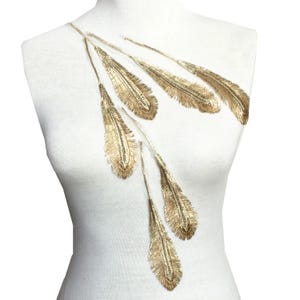 May include: A white garment with a gold embroidered feather design. The feathers are arranged in a cascading pattern, with detailed stitching and a metallic sheen. The design is displayed on a mannequin.
