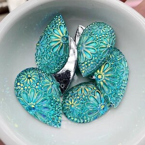 Pode incluir: Enfeites em forma de gota de água turquesa e amarela com desenhos florais. Os enfeites estão dispostos numa tigela branca. As gotas têm uma textura brilhante e também está presente uma peça prateada metálica.